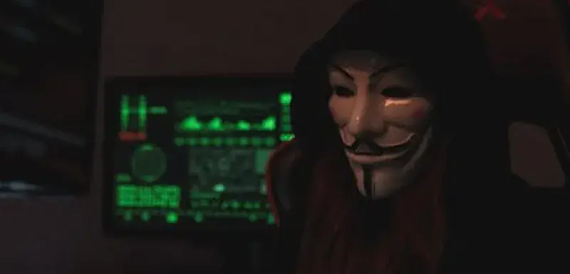 Personne anonyme portant un masque Guy Fawkes travaillant sur un ordinateur dans une pièce sombre, évoquant des activités cybernétiques