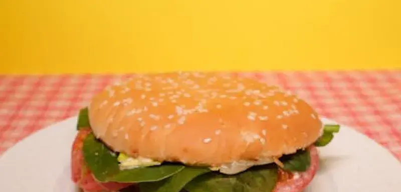 Delicious burger with green salad leaves and tomatoes with sauce placed on white plate on table near yellow wall