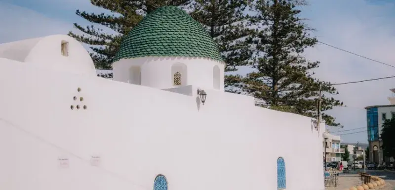 A whitewashed Mediterranean building with a green dome, surrounded by trees, and under a clear blue sky.