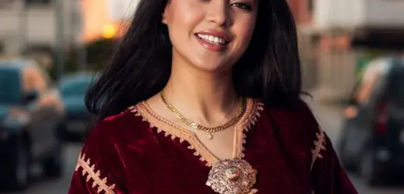 Elegant woman in a red velvet dress smiling on a street during sunset.