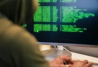 Personne en sweat à capuche tapant du code sur un écran d'ordinateur, représentant une scène de piratage informatique