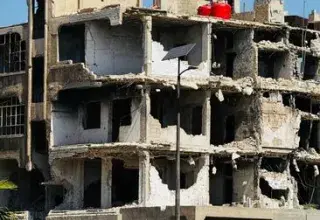 Desolate war-damaged building in Homs, Syria, with a truck in foreground.
