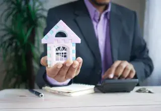 man holding model house at desk with calculator